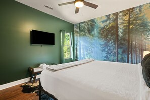 Un dormitorio con cama, televisor de pantalla plana, ventilador de techo, ventana con cortinas y un mural de un bosque en la pared.