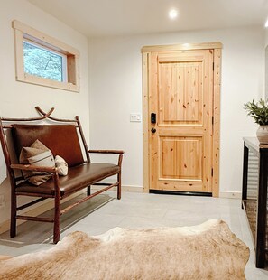 Una habitación con una puerta de madera, un banco de cuero con cojines, una alfombra y una planta pequeña sobre una mesa.