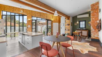 Une cuisine moderne dotée d’un îlot central, de poutres en bois, de grandes fenêtres et d’un coin repas avec des chaises.