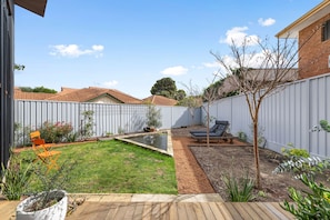 Un patio trasero con césped, un estanque pequeño, una terraza de madera y un área cercada con un árbol y un banco.