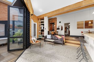 Um interior moderno com entrada por porta de vidro, área de estar com sofá e poltronas, cozinha com mesa de jantar e teto de madeira.