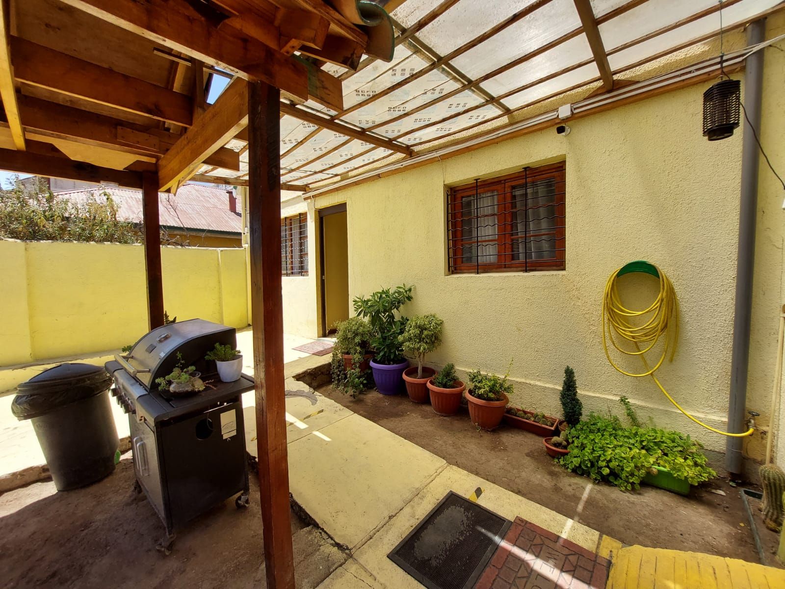 A covered patio area with a grill, potted plants, and a hose.