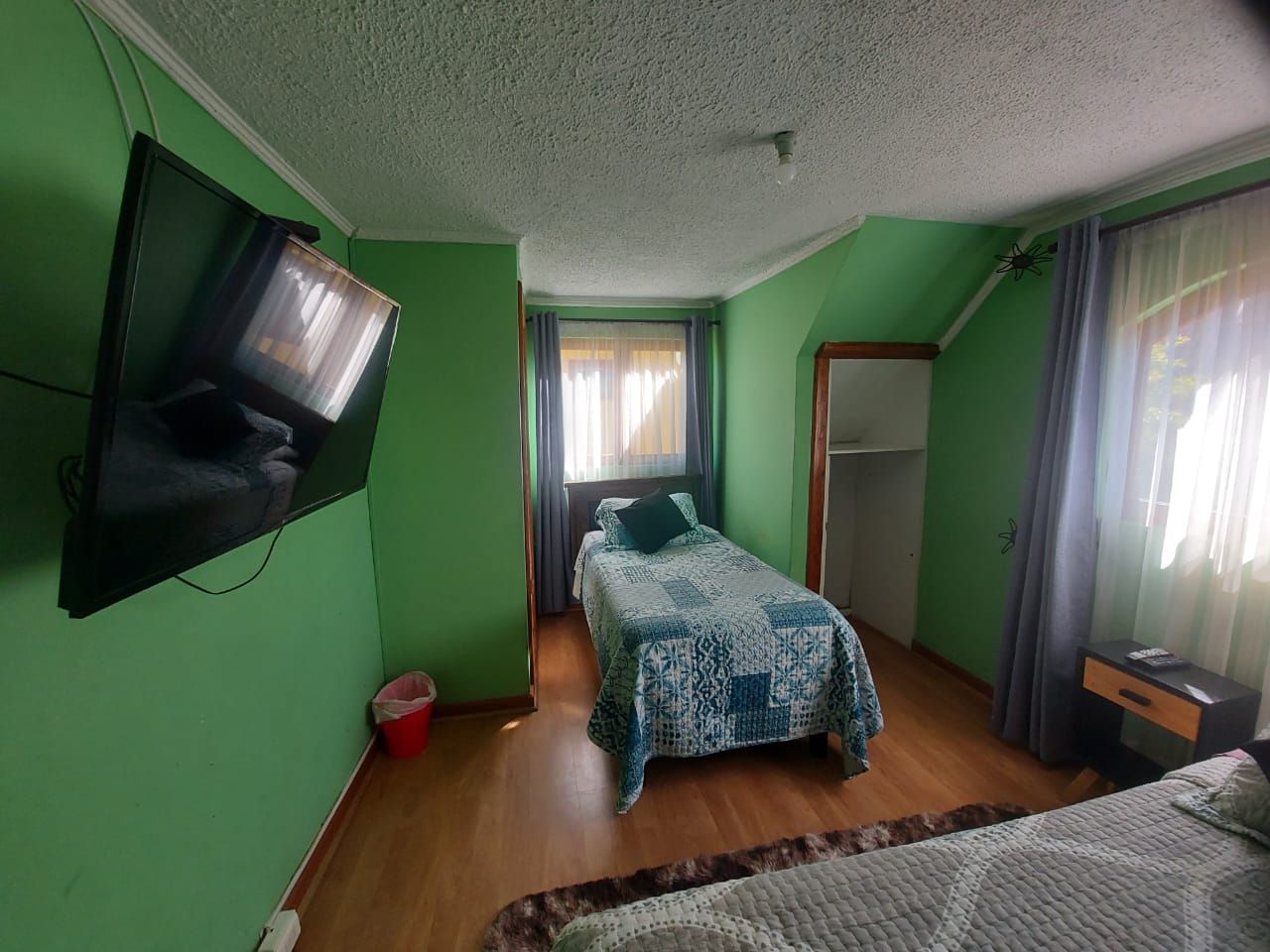 Ein Zimmer mit einem Bett, einem an der Wand montierten Fernseher, einem Fenster mit Vorhängen und einer Tür.