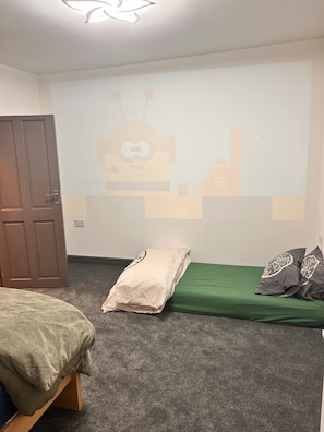 Une chambre à coucher avec un lit, un matelas vert, un oreiller, un projecteur fixé au mur qui diffuse un dessin animé, et une porte.