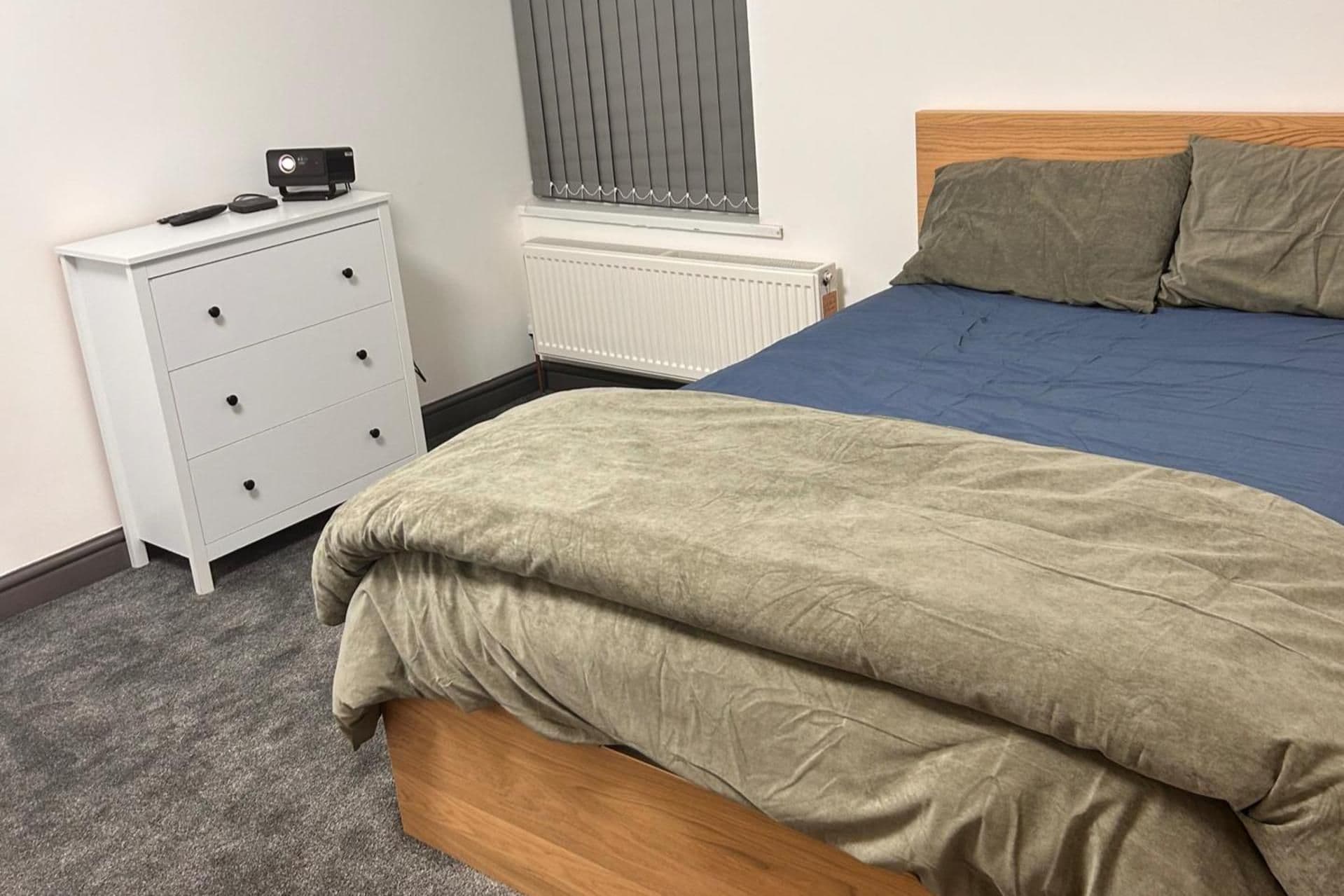 Un dormitorio con cama, cómoda, radiador y ventana con persianas.