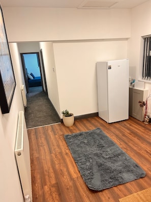 Una cocina compacta con un refrigerador blanco, una pequeña planta en maceta y una alfombra gris sobre piso de madera.