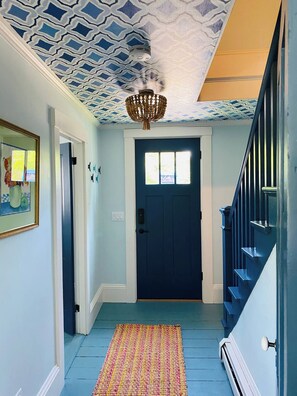 Un couloir avec un plafond orné de motifs, une porte bleu foncé, un escalier et un tapis coloré.