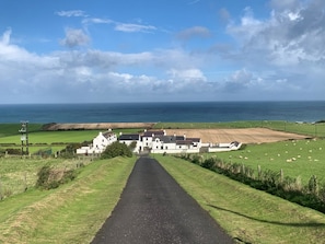 A rural road leading to a white building with multiple rooms, surrounded by fields and a view of the sea. - Cosy Cottage on the North Antrim Coast of Northern Ireland near Ballintoy. (Ballintoy)