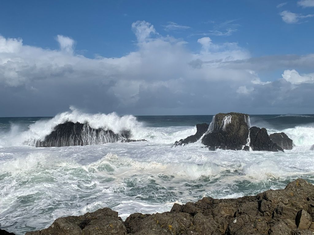 A coastal scene with waves crashing against rocks under a partly cloudy sky.