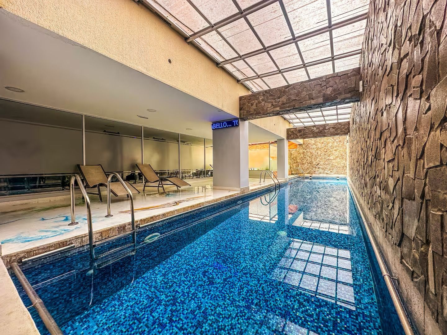An indoor swimming pool with clear blue water, lounge chairs, and a stone wall.