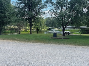 A park with a pond, trees, a trash can, and a picnic table.