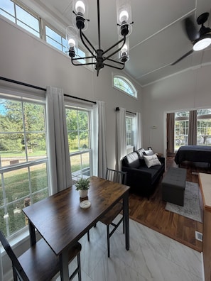 A spacious living room with a large window, a black sofa, a wooden dining table, and a ceiling fan.