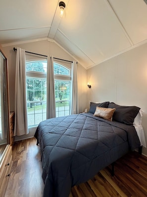 A bedroom with a large bed, a window with curtains, a bedside table, and a wall-mounted light.