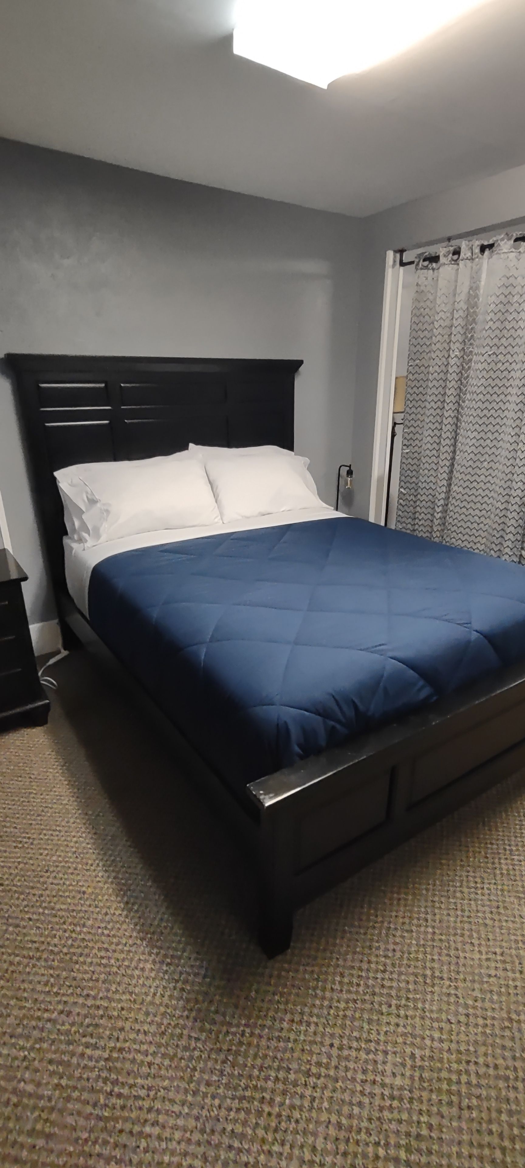 A bed with a dark headboard, white pillows, and a blue bedspread in a room with a patterned carpet.