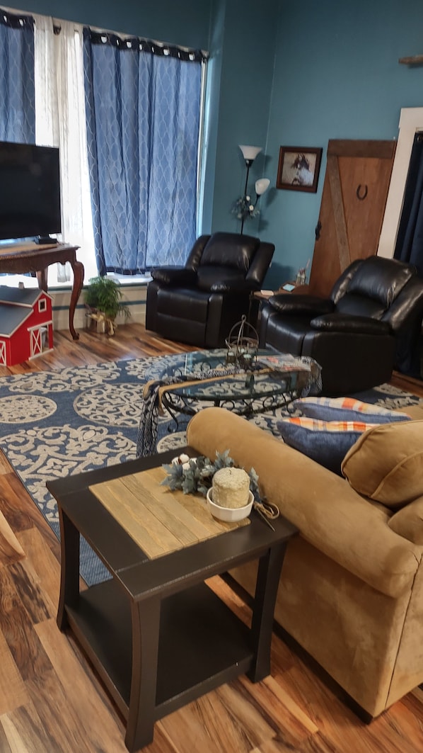 Una sala de estar con un sofá, dos sillones, una mesa de centro, un televisor y una alfombra estampada.