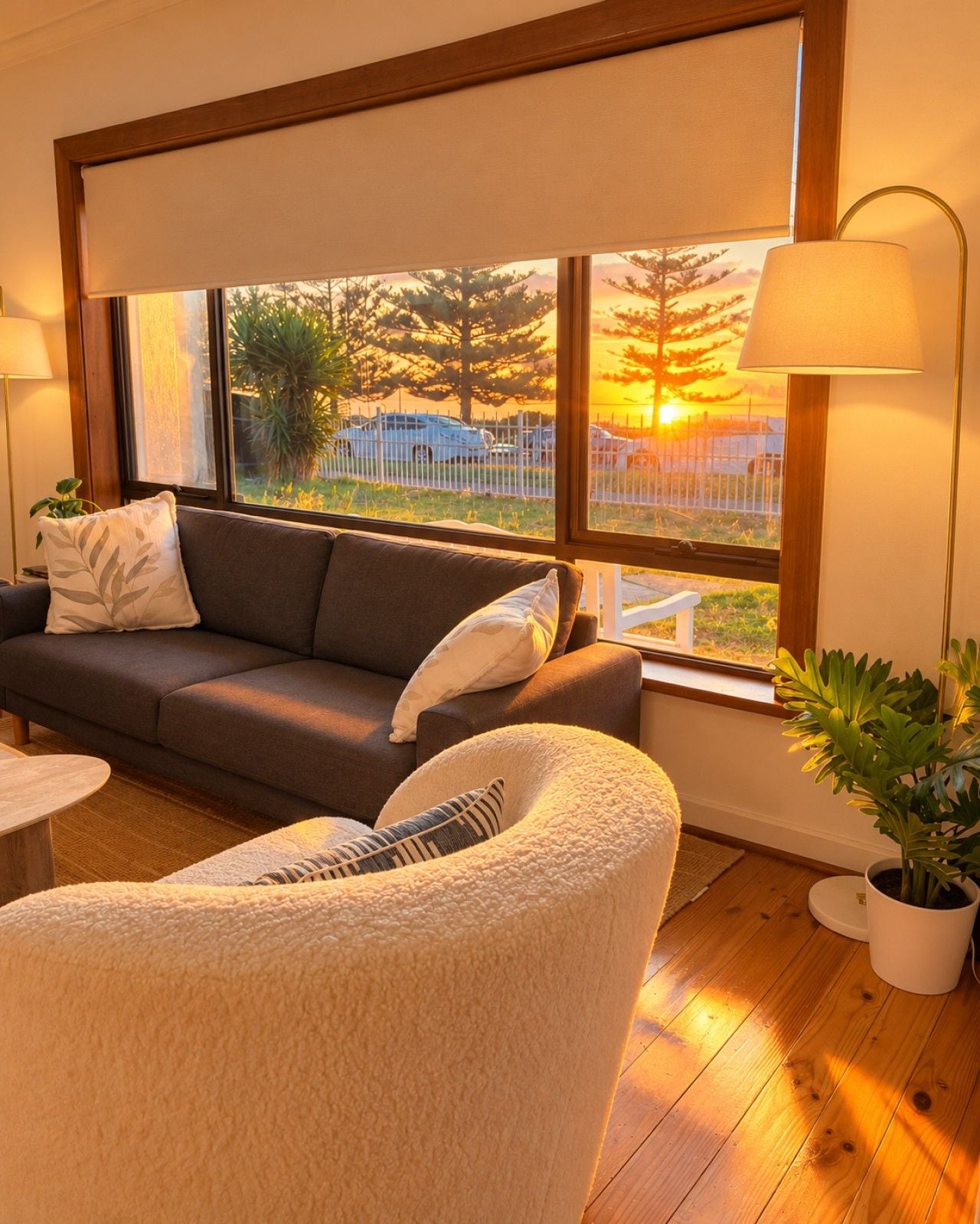 A cozy living room with a sofa, a chair, a lamp, and a view of a sunset through large windows.