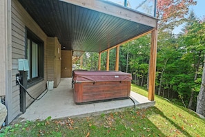 A wooden cabin with a covered porch, a hot tub, and a view of a forested area.