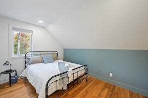 A bedroom with a bed, a window with a view of trees, a bedside table with a lamp, and a blue wall.