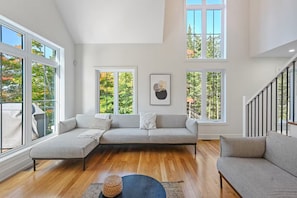 A spacious living room with a sectional sofa, a round ottoman, and large windows offering views of trees.