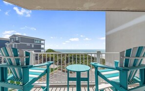 Een balkon met turquoise stoelen en een kleine tafel, met uitzicht op een parkeerplaats en een gebouw met een bord.