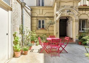 A courtyard with a red metal table and chairs, surrounded by potted plants and a building with a balcony. - Buttes-Chaumont · 19th · Modern & Well-Equipped Studio – by Mey Agency (Paris)
