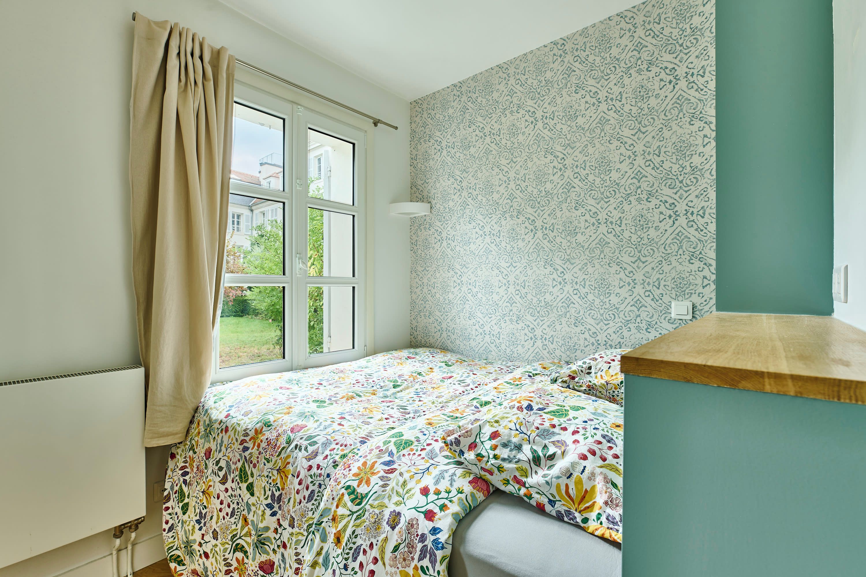 Une chambre à coucher avec un lit, une fenêtre avec des rideaux, un papier peint à motifs et un interrupteur sur le mur.