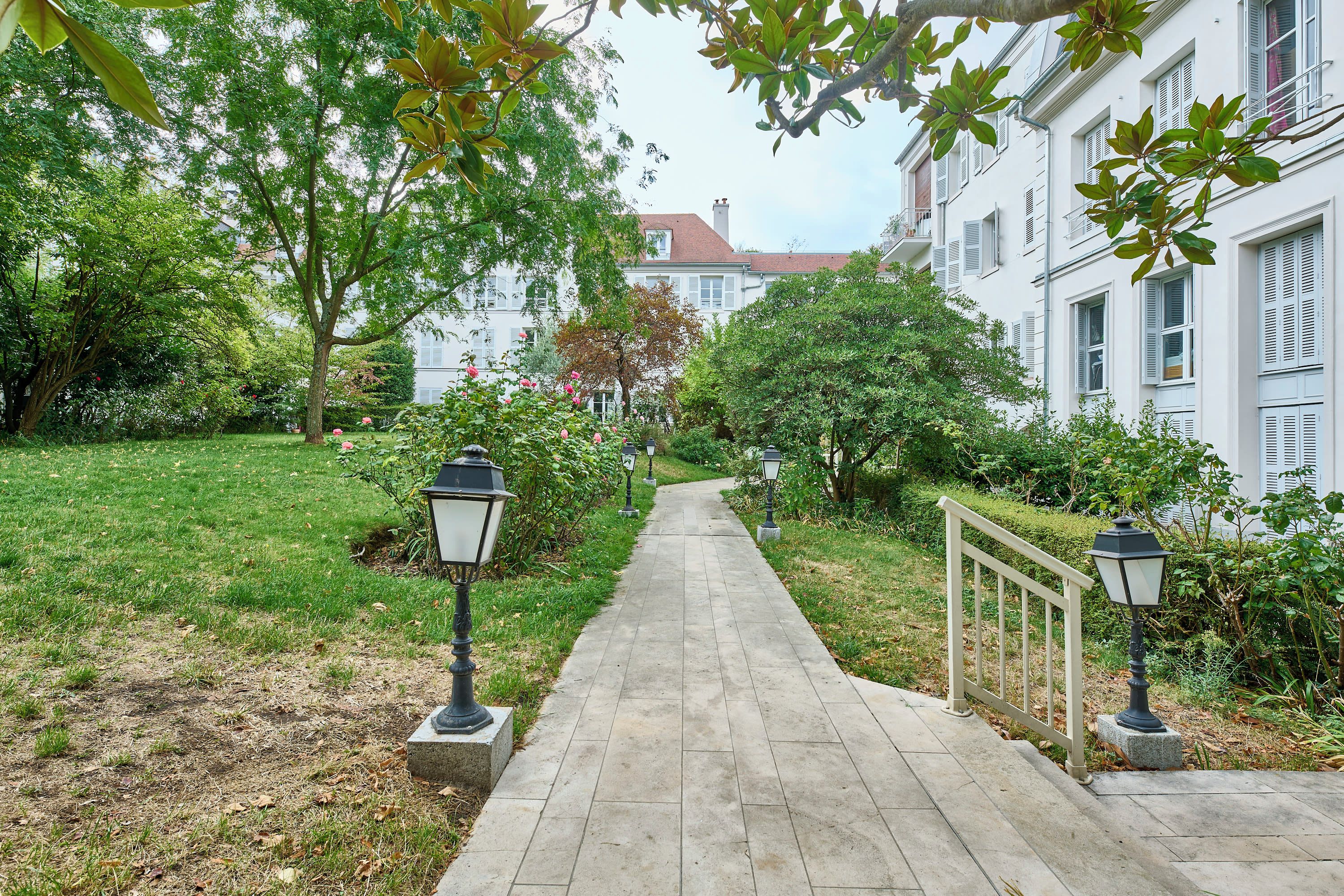 Un chemin qui mène à une rangée de bâtiments blancs avec des volets, entourés de verdure et éclairés par des lampadaires.