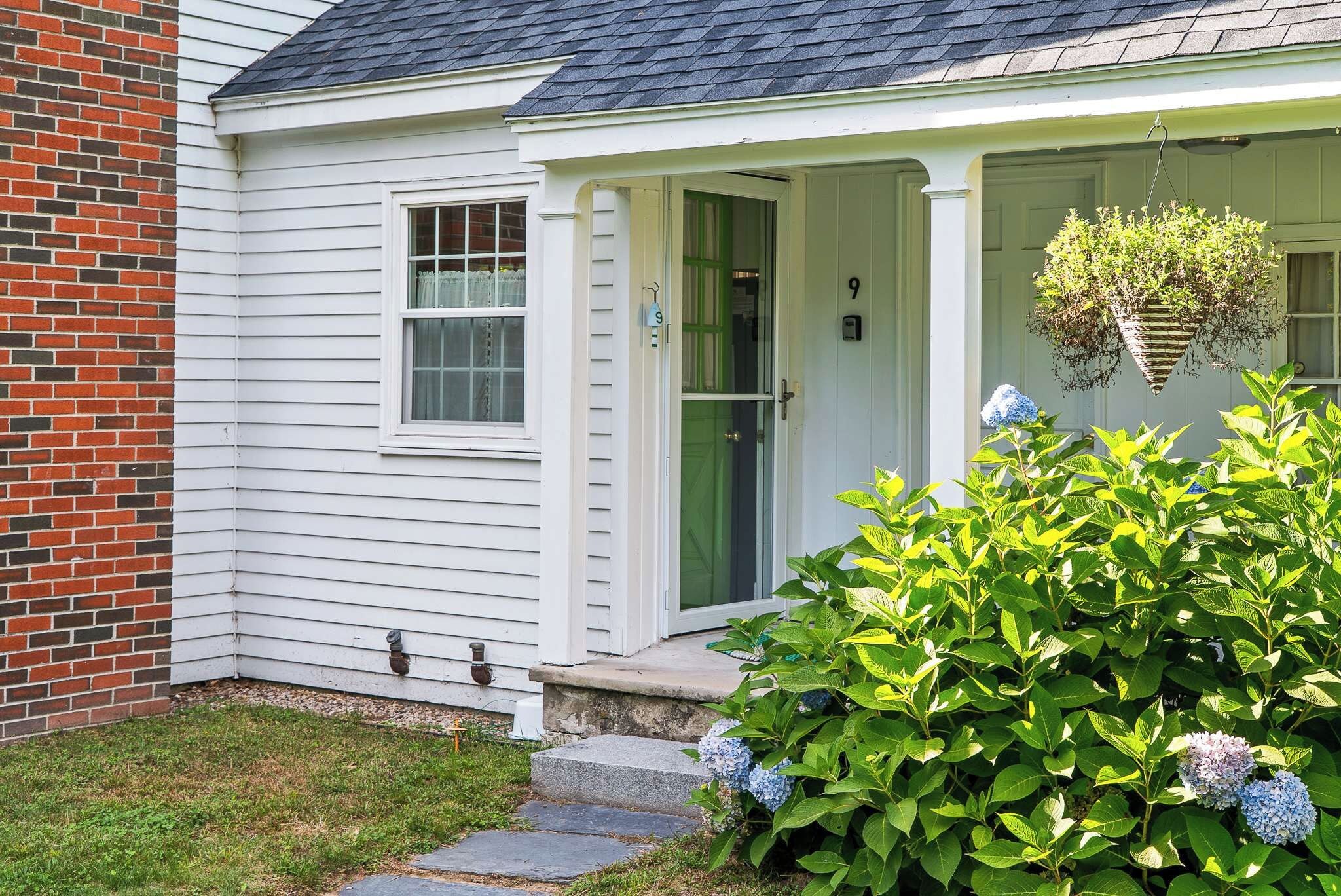 A house with a white exterior, a brick section on the left, a green door, and a small porch with a hanging plant.