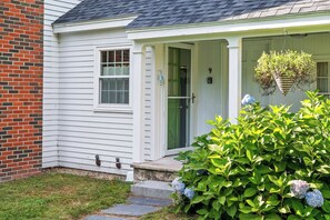 A house with a white exterior, a brick section on the left, a green door, and a small porch with a hanging plant. - A Wave From It All with Hot tub (Kennebunk)