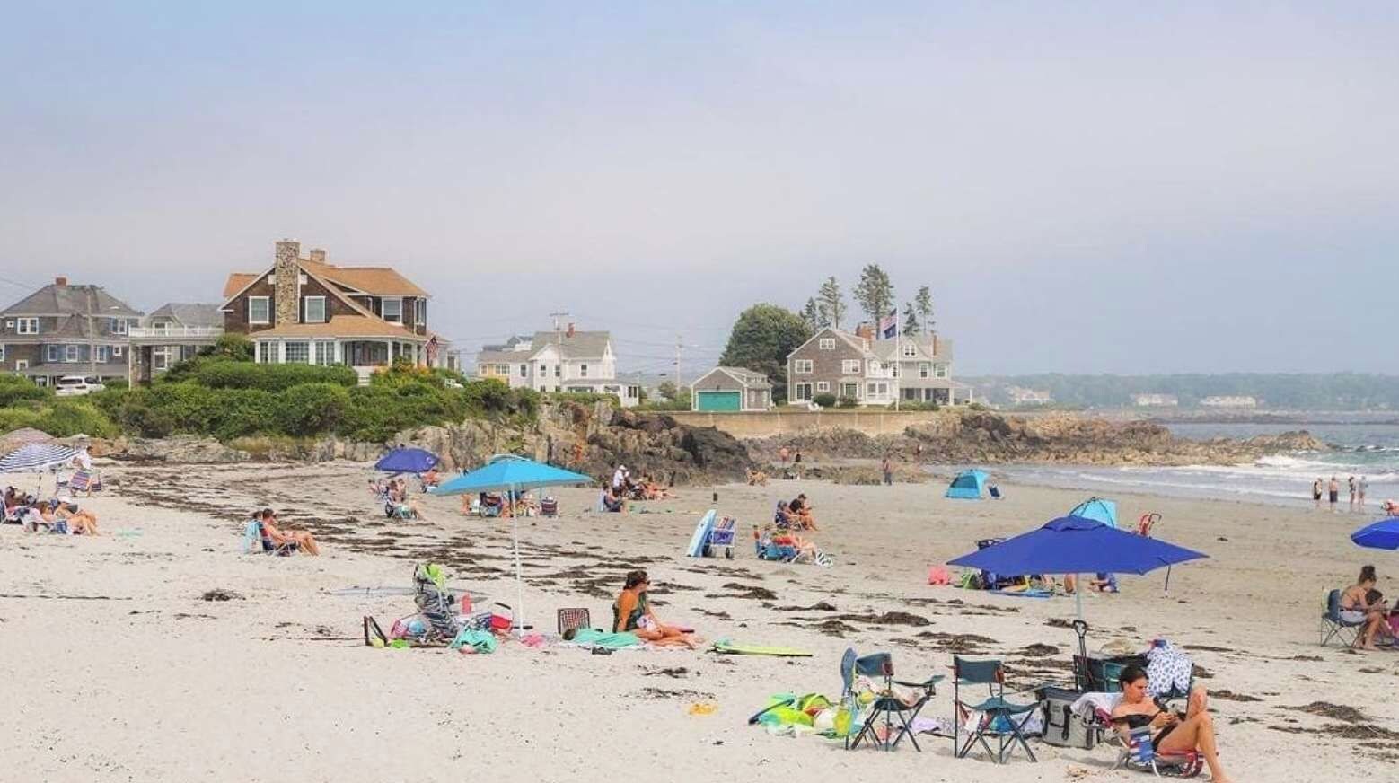 A beach with people, umbrellas, and houses in the background.