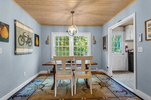A dining area with a wooden table and chairs, a chandelier, and a window with a view of greenery. - A Wave From It All with Hot tub (Kennebunk)