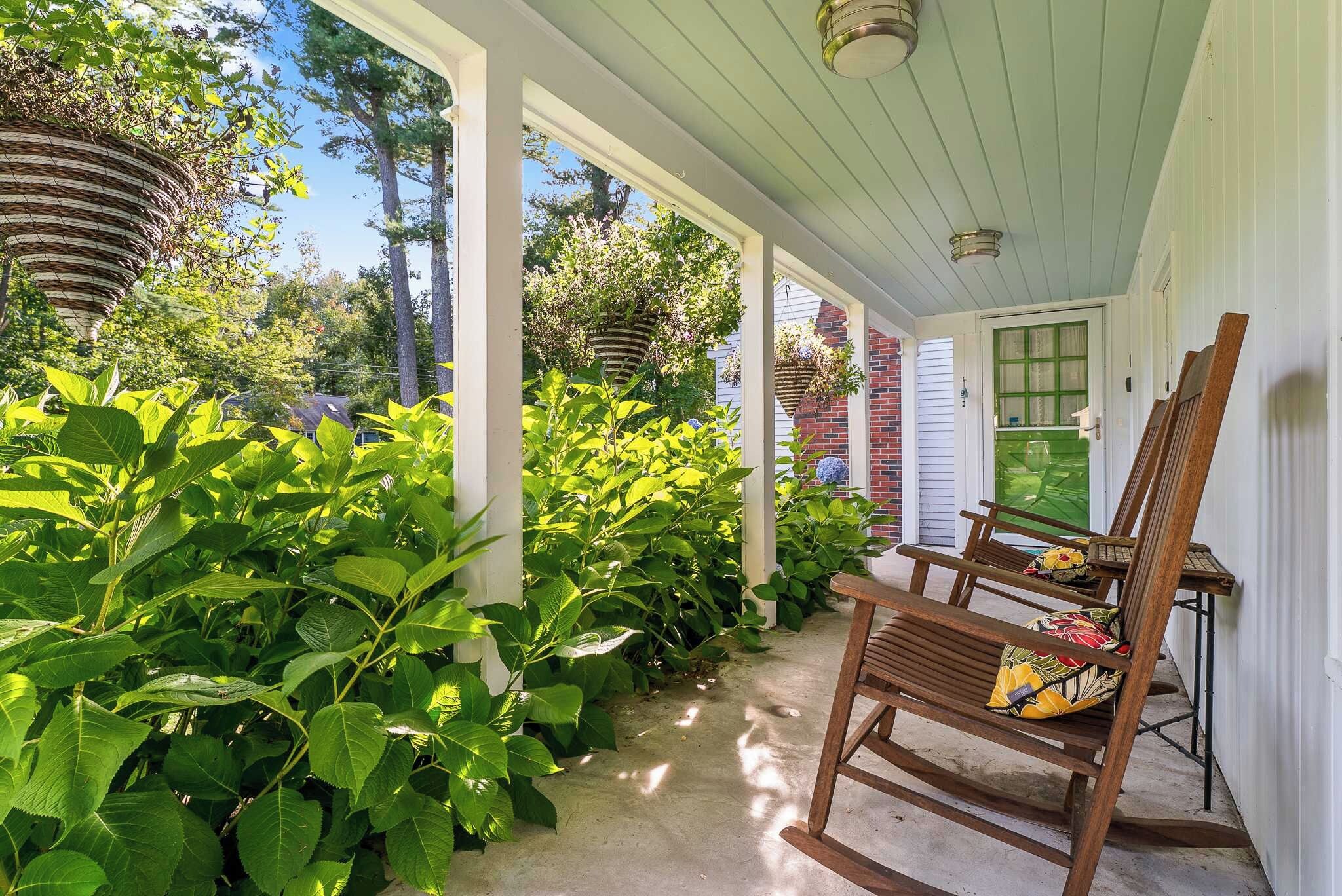 A covered porch with a wooden rocking chair and a table, surrounded by lush greenery.
