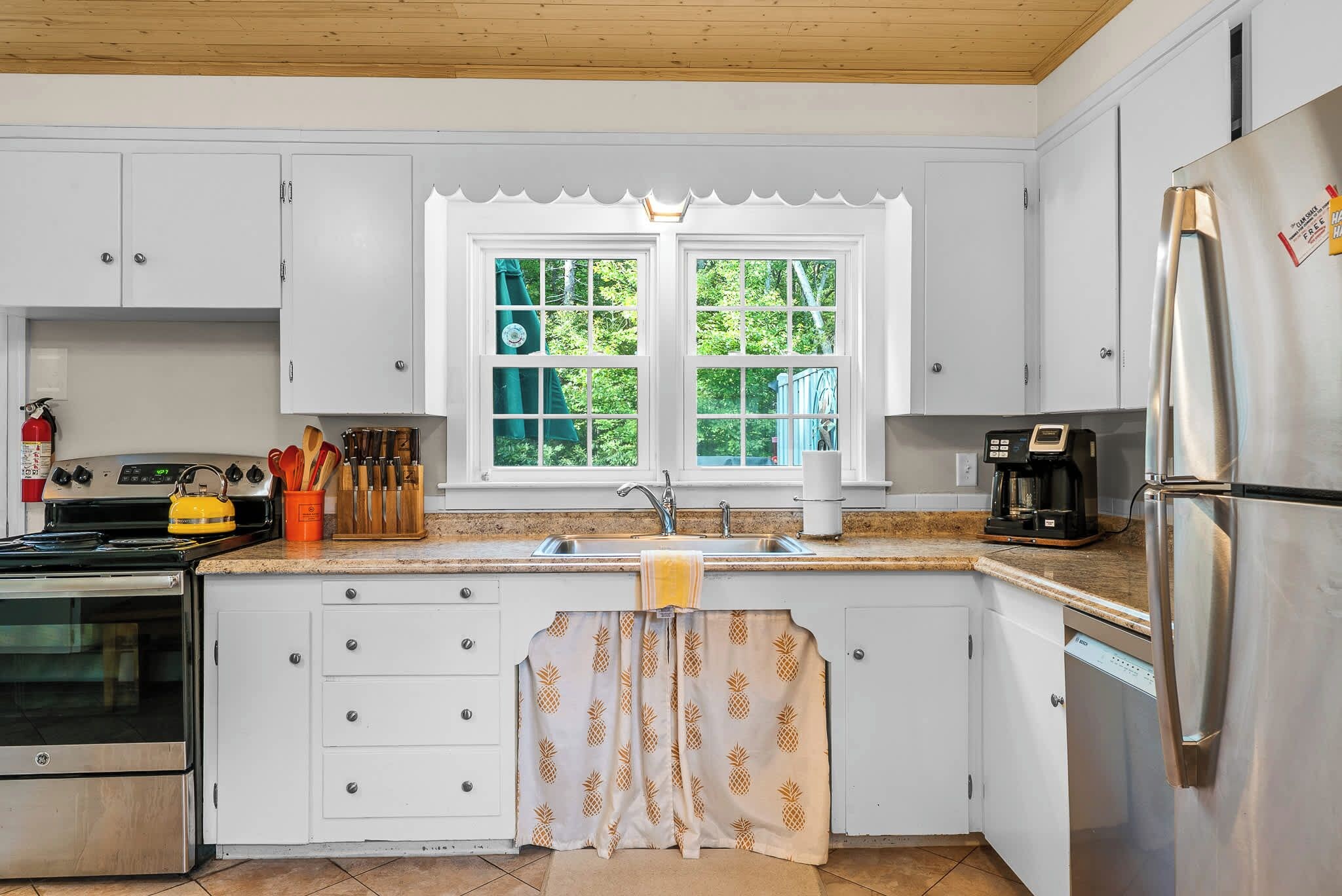 Una cocina con armarios blancos, un refrigerador de acero inoxidable, una estufa y un ventanal con vistas a la vegetación.