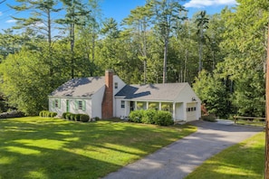 Une maison de plain-pied avec une cheminée, entourée d’une pelouse bien entretenue et d’arbres touffus.
