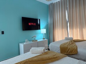 Ein Hotelzimmer mit einem Bett, einem Nachttisch mit Lampe, einem Fernseher, der 'NETFLIX' anzeigt, und einem Fenster mit Vorhängen.