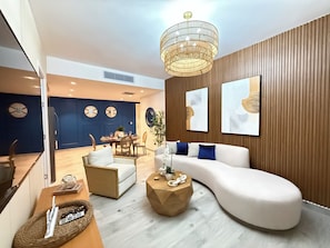 A modern living room with a curved white sofa, a wooden coffee table, and a dining area with a blue wall and clocks. - Your Oasis in Punta Cana: 2-Bedroom Apartment for 6 People with a Pool (Punta Cana)