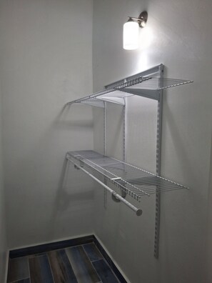 A corner with two metal wire shelves mounted on a wall, a metal rack with a pull handle, and a light fixture above the top shelf. - 2 Walking to the beach (Noord)