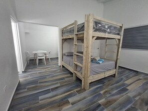 A bunk bed with a wooden frame and a mattress, a small white table with a chair, and a tiled floor. - 2 Walking to the beach (Noord)