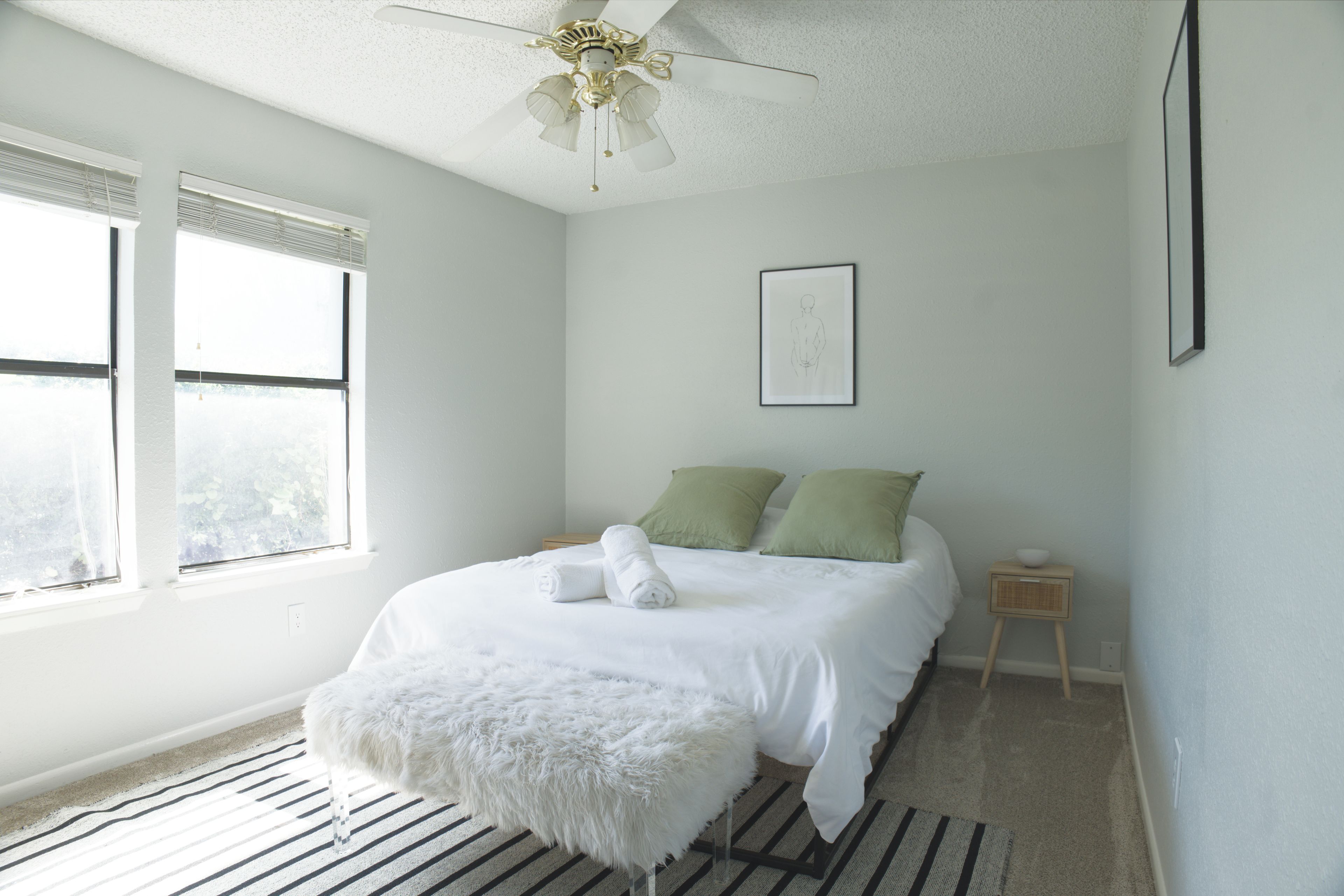 A bedroom with a bed, pillows, a bedside table, a small stool, a framed picture on the wall, and a ceiling fan.
