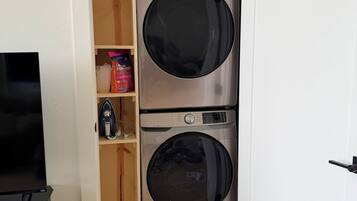 A laundry room with two stacked front-loading washing machines, a shelf with laundry detergent, and a television on a stand.