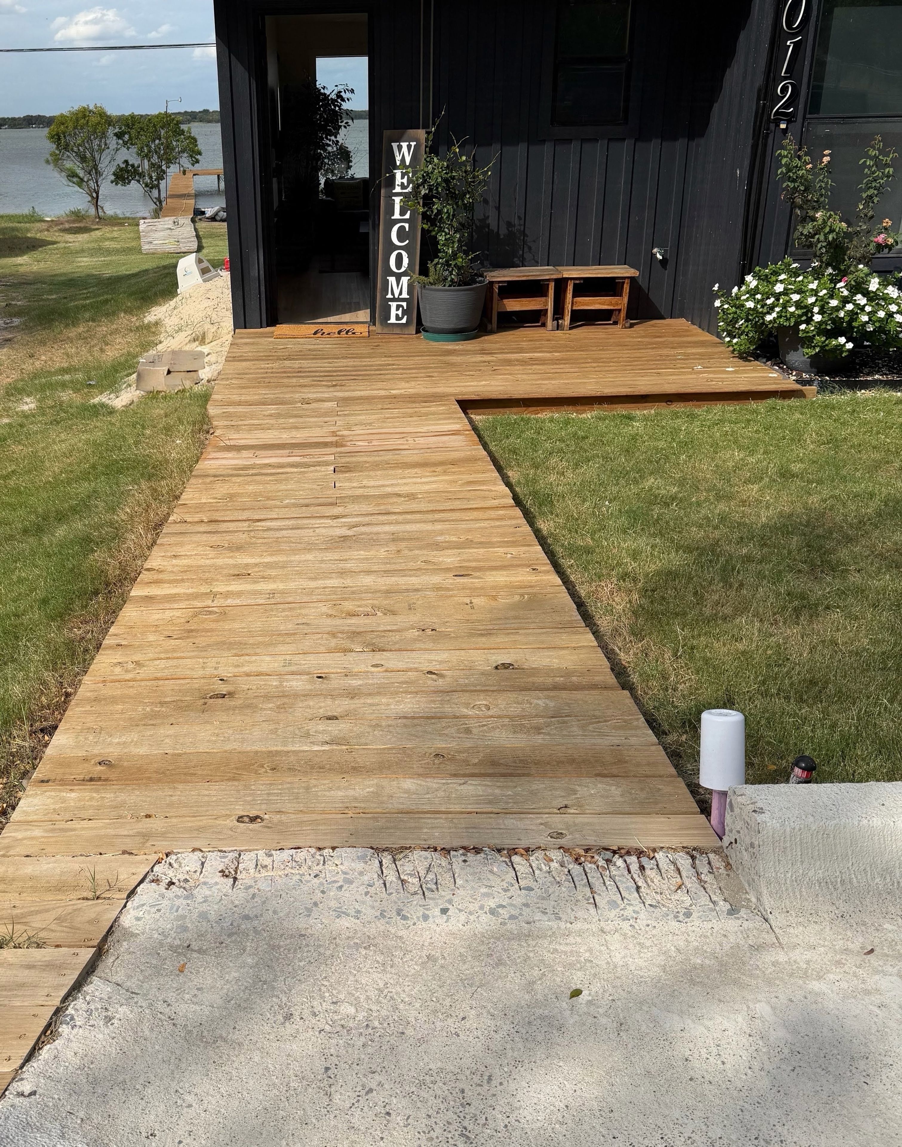 En træterrasse fører til en mørk bygning med et 'WELCOME'-skilt, et lille udendørs siddeområde og udsigt til vandet.