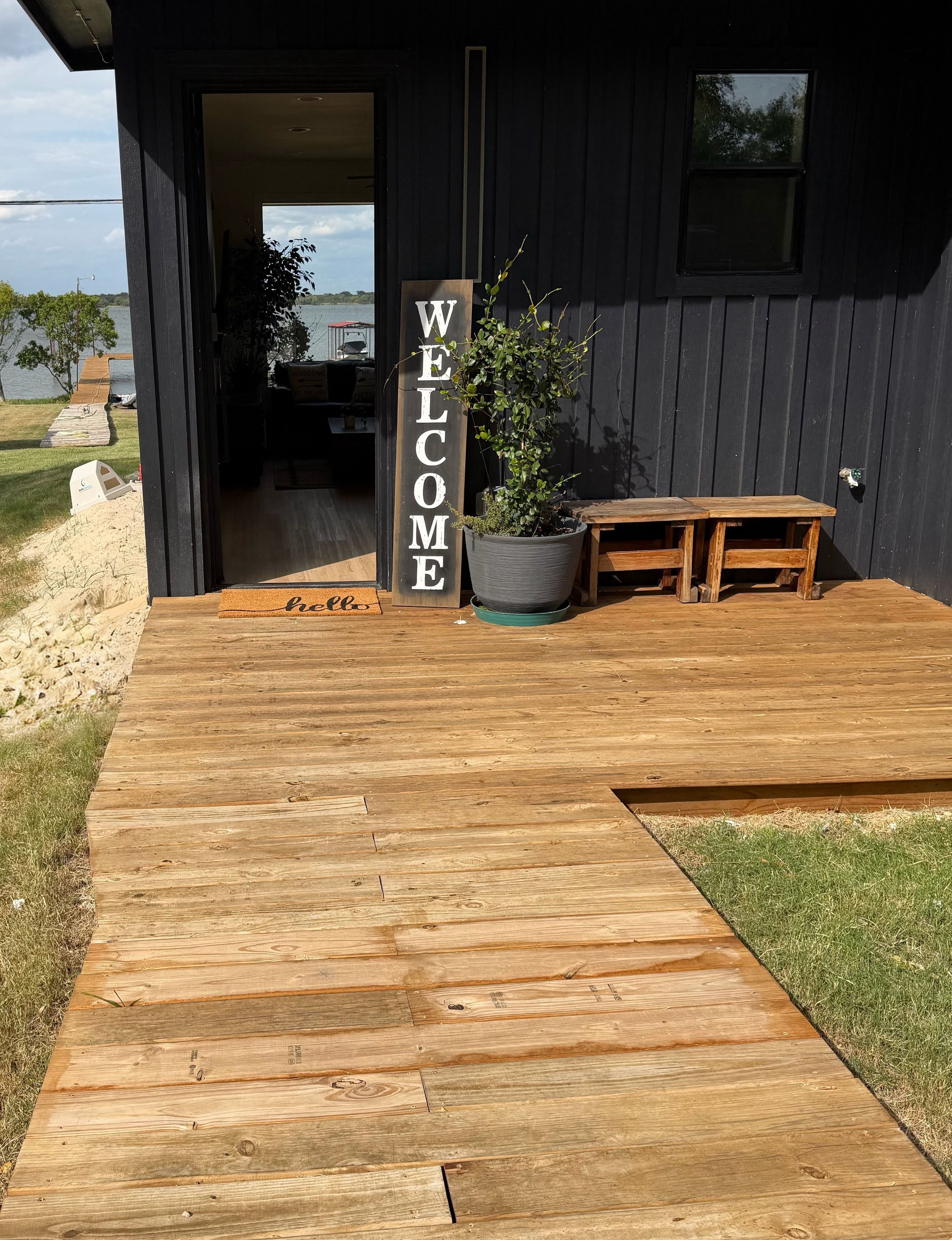Una terraza de madera que lleva a un edificio de color oscuro con un letrero que dice 'WELCOME', una planta en maceta y un banco.