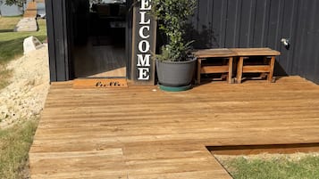 Una terraza de madera que lleva a un edificio de color oscuro con un letrero que dice 'WELCOME', una planta en maceta y un banco.