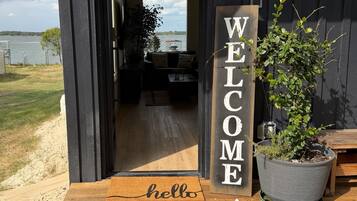 A wooden door with a 'WELCOME' sign, a doormat with 'hello' text, and a potted plant on a wooden deck.