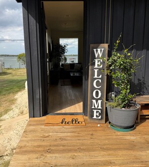 A wooden door with a 'WELCOME' sign, a doormat with 'hello' text, and a potted plant on a wooden deck.