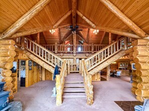 A log cabin interior with a central staircase, wooden walls, and a ceiling fan. - Historic 5-bedroom log home in Choteau perfect for families or large groups (Choteau)