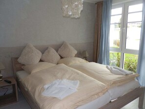 Un lit double avec un cadre en bois, une table de chevet avec une lampe, et une vue sur la verdure par la fenêtre.
