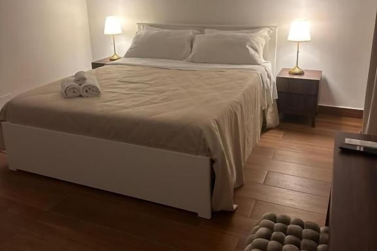 A bedroom with a bed, bedside lamps, a wooden floor, and a bench.