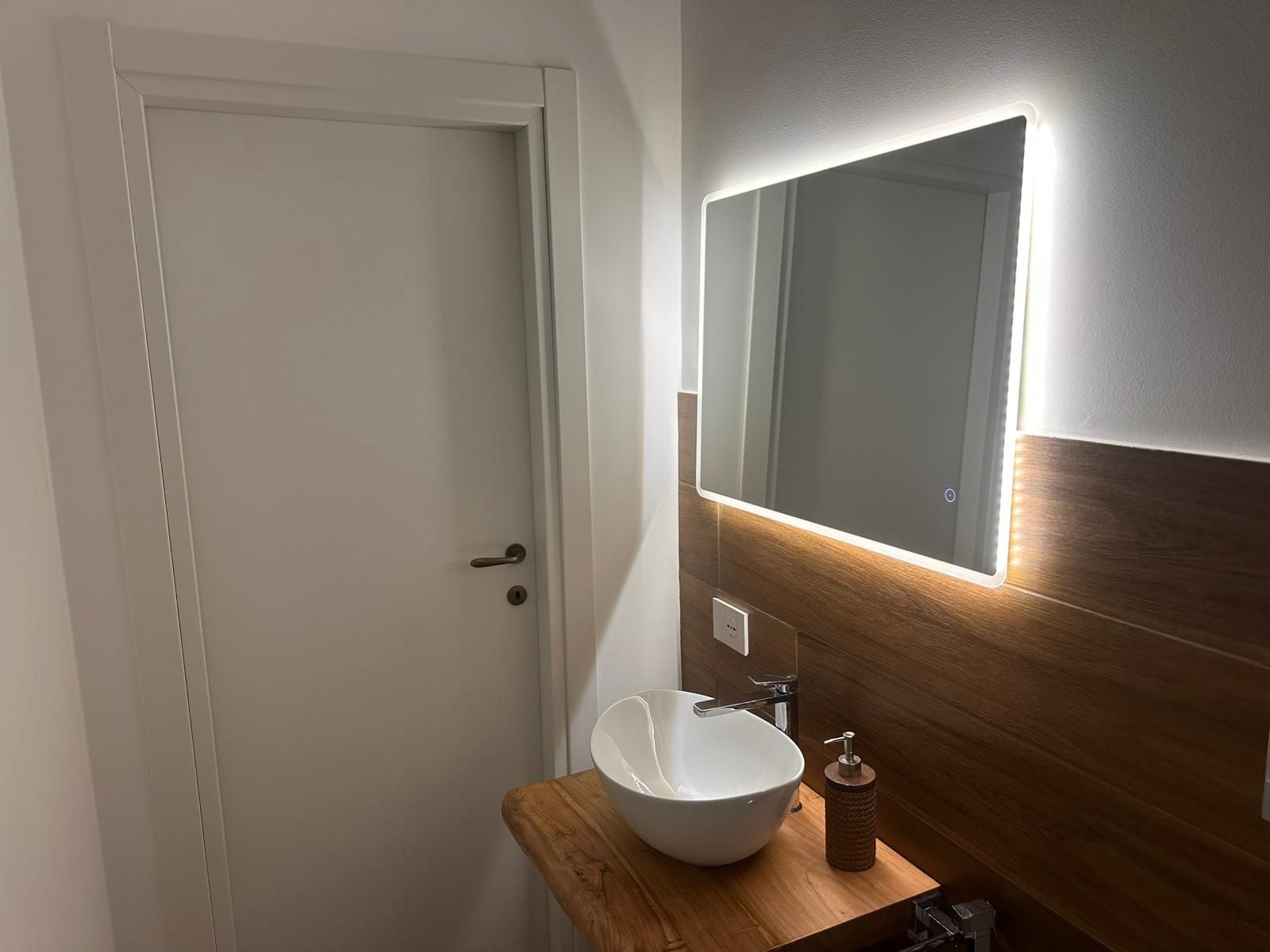 A bathroom with a white door, a wooden vanity, a white sink, a mirror with integrated lighting, and a soap dispenser.
