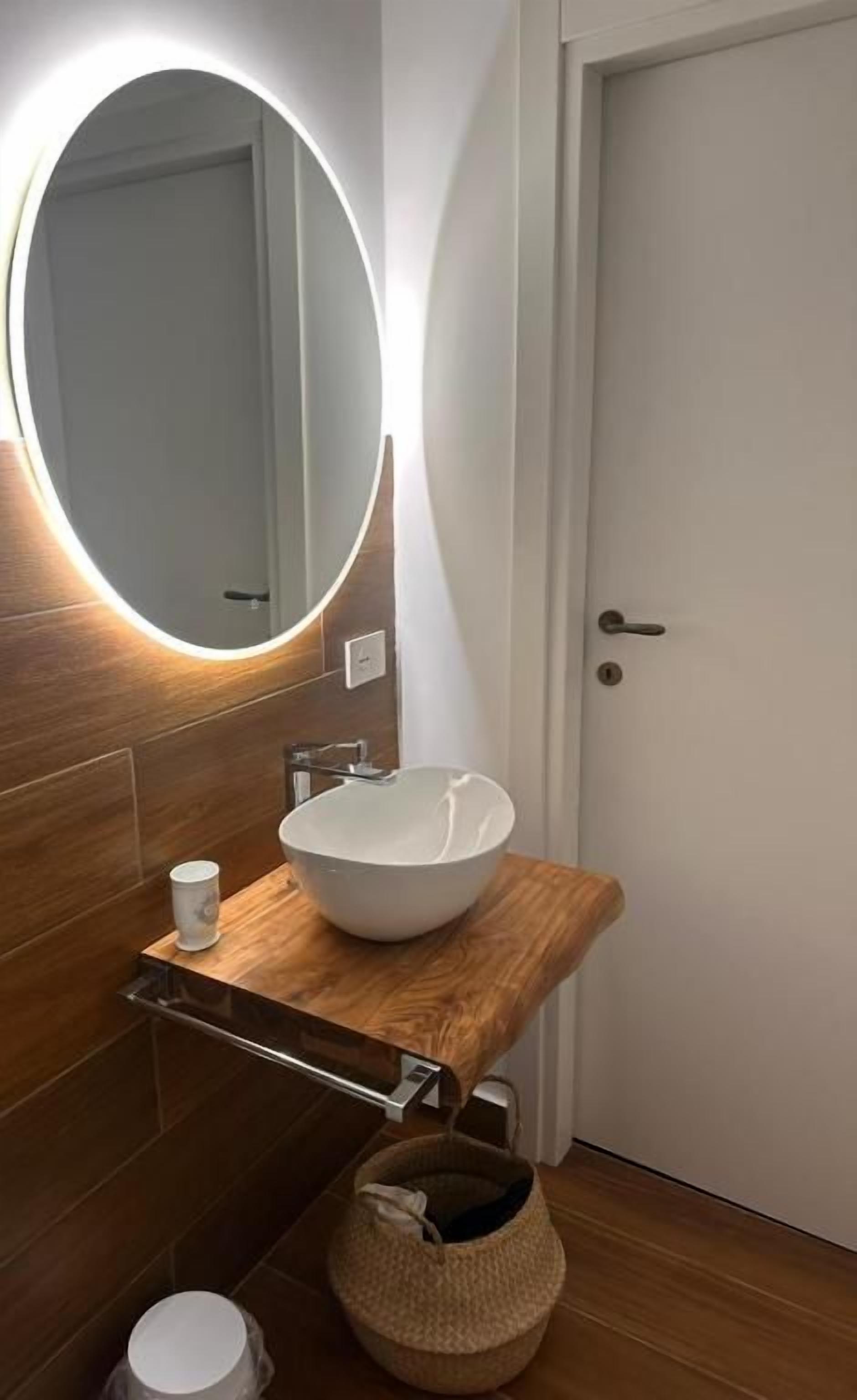 Un baño con tocador de madera, un lavabo blanco, un espejo redondo con iluminación, una taza blanca pequeña y una cesta tejida.
